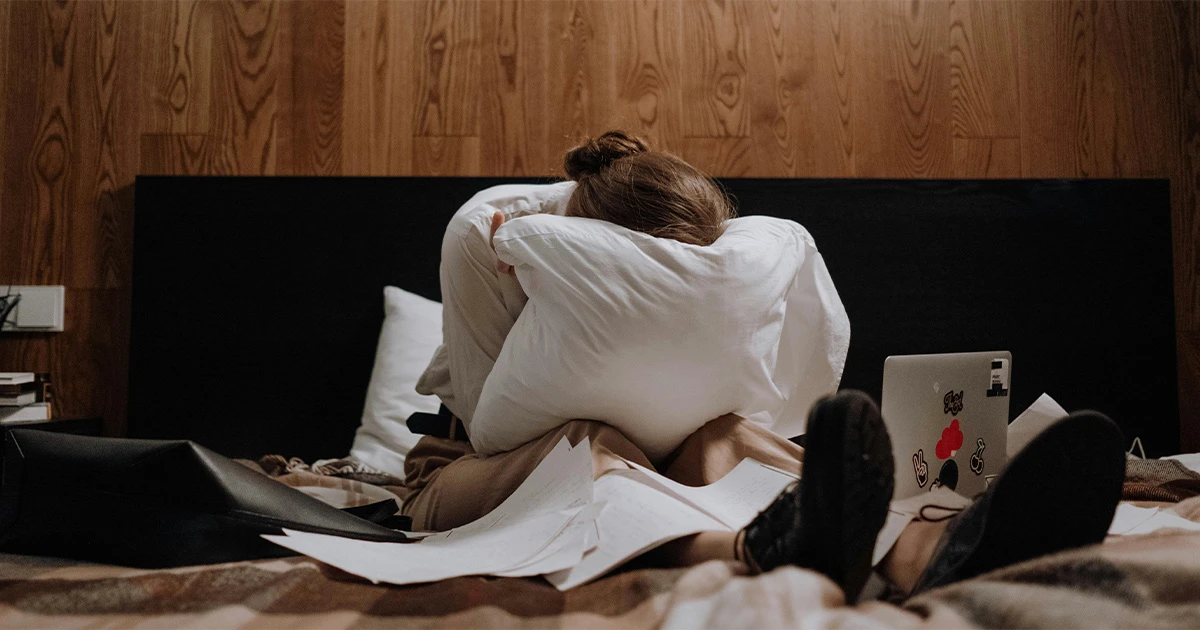 A lady dipping her face onto a white pillow in front of her laptop due to overwhelming negative feelings displays one of the 3 harsh facts about long-distance relationships