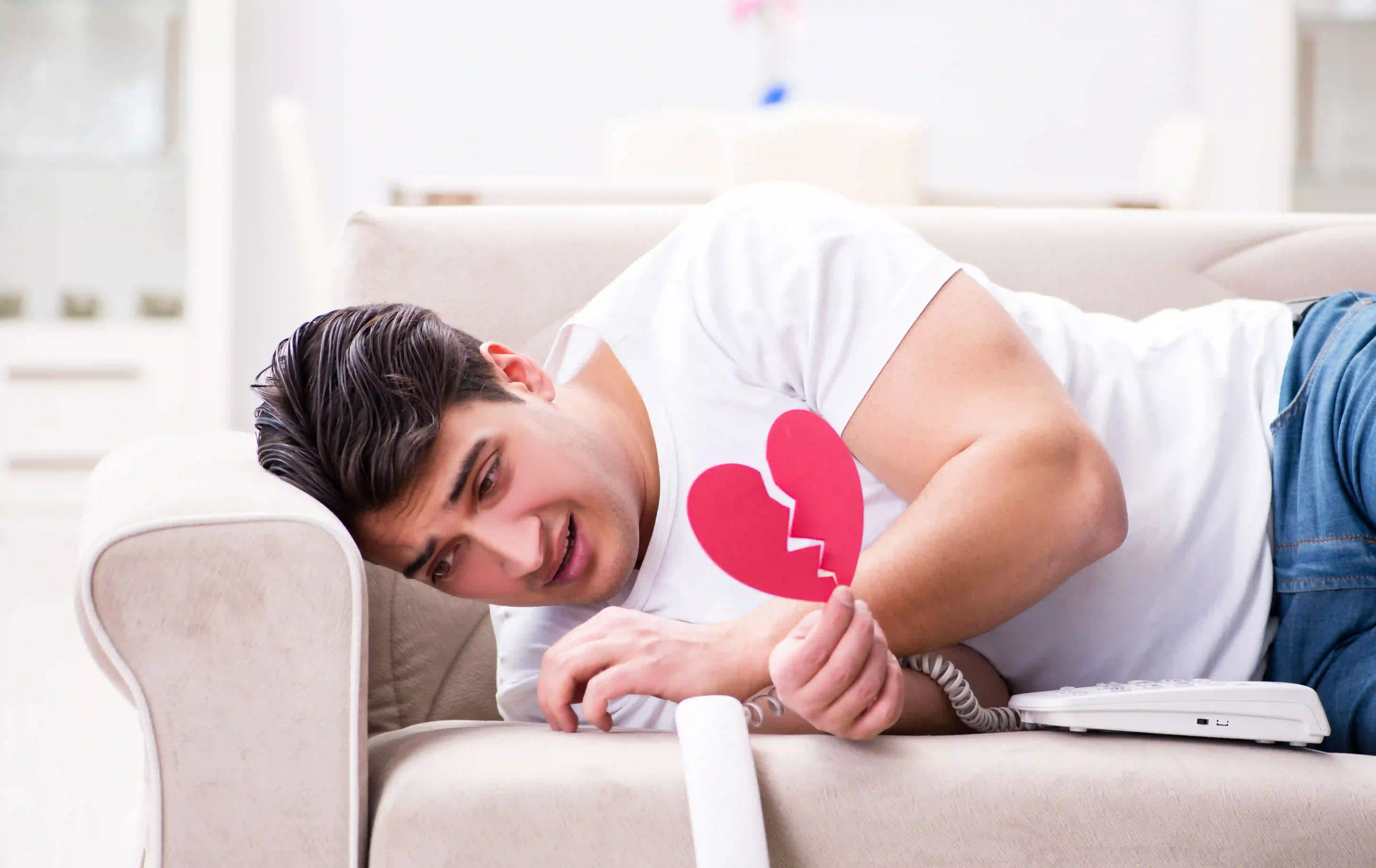 A man lying on a couch by the telephone and holding paper shaped like a broken heart