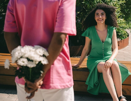 A guy in a pink shirt with a hidden bouquet of flowers at his back and facing a smiling lady he likes as he attempts to determine how to initiate a conversation with her”