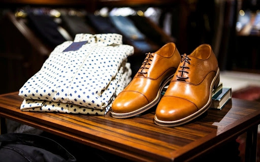 Two folded shirts and a pair of brown shoes on a wooden table