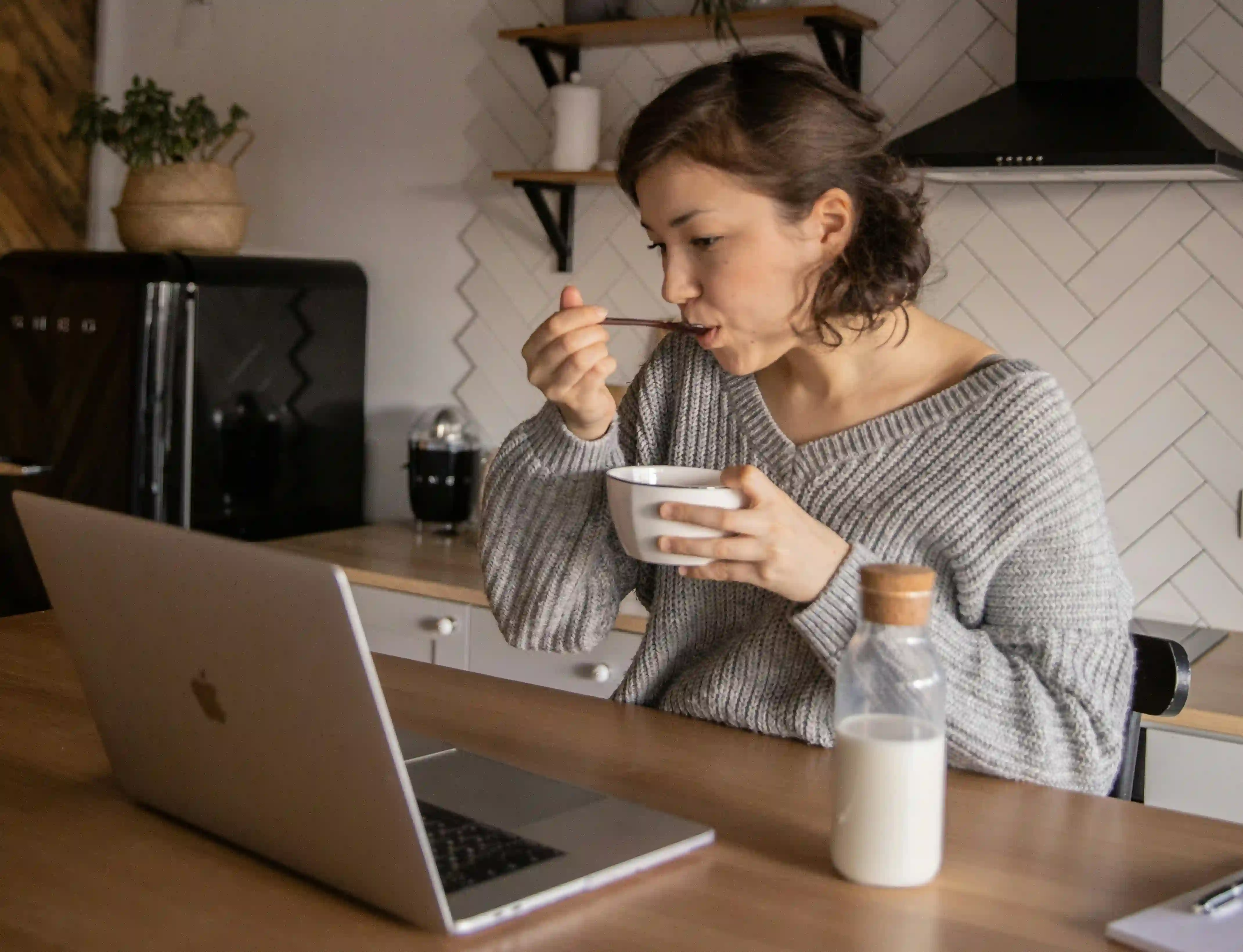 A woman in a gray sweatshirt eating food from a bowl while having a video call using her laptop