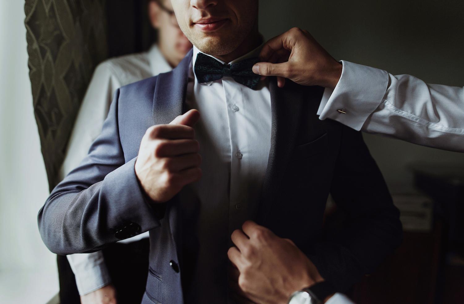 A man wearing a dark suit while someone fixes his bow tie